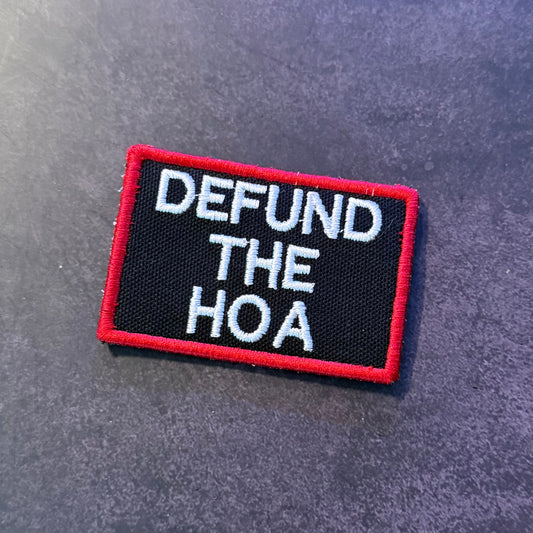 A rectangular embroidered patch with a black background, red border, and white text reading 'DEFUND THE HOA', placed on a dark textured surface.