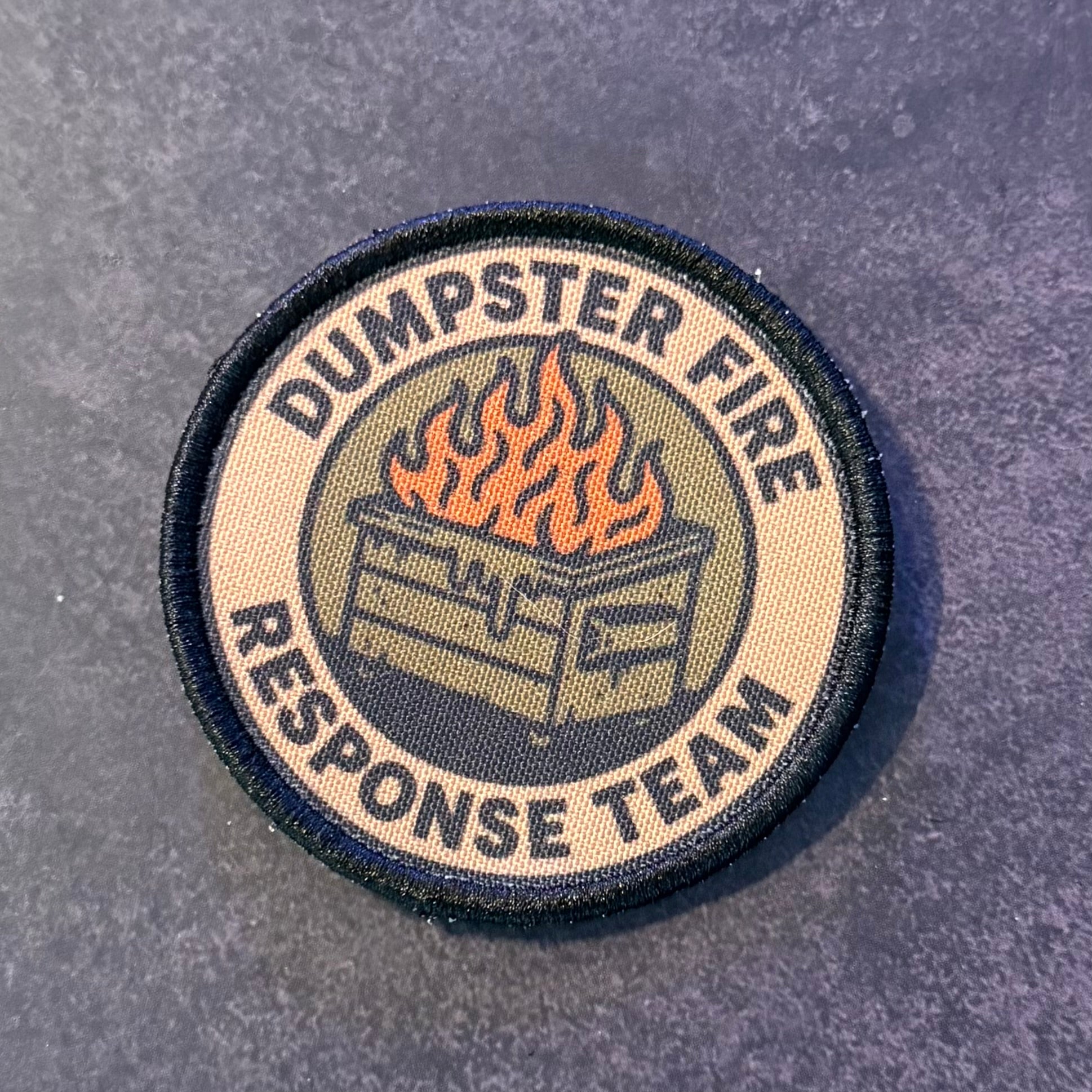 A circular embroidered patch reading 'DUMPSTER FIRE RESPONSE TEAM' with a central fire-and-dumpster graphic, shown on a dark textured surface.