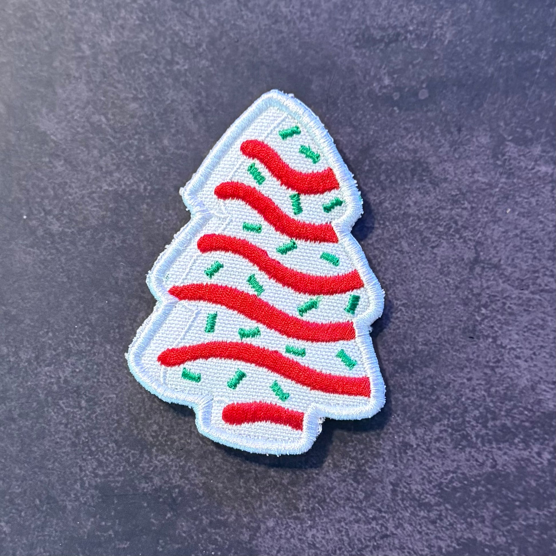 An embroidered Christmas tree patch with a white background, red wavy stripes, and green sprinkle details, shown on a dark textured surface.