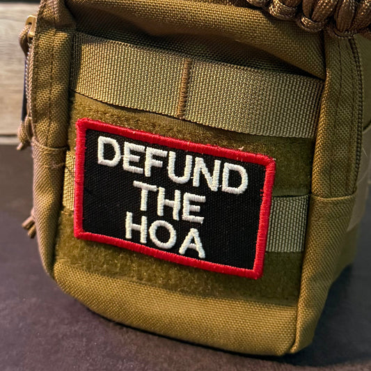 An olive drab tactical pouch with a black and red embroidered patch reading 'DEFUND THE HOA' in white letters, featuring a paracord strap and shown against a rustic wood background.