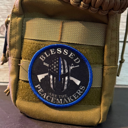 A tan tactical pouch with a thick woven strap, featuring a circular patch that reads 'BLESSED ARE THE PEACEMAKERS' in white text with a blue border. The pouch is set against a light wood plank background.