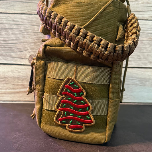 A tactical utility pouch in olive drab canvas with a brown paracord shoulder strap and a red and green embroidered Christmas tree patch on the front, set against a light wood plank background.