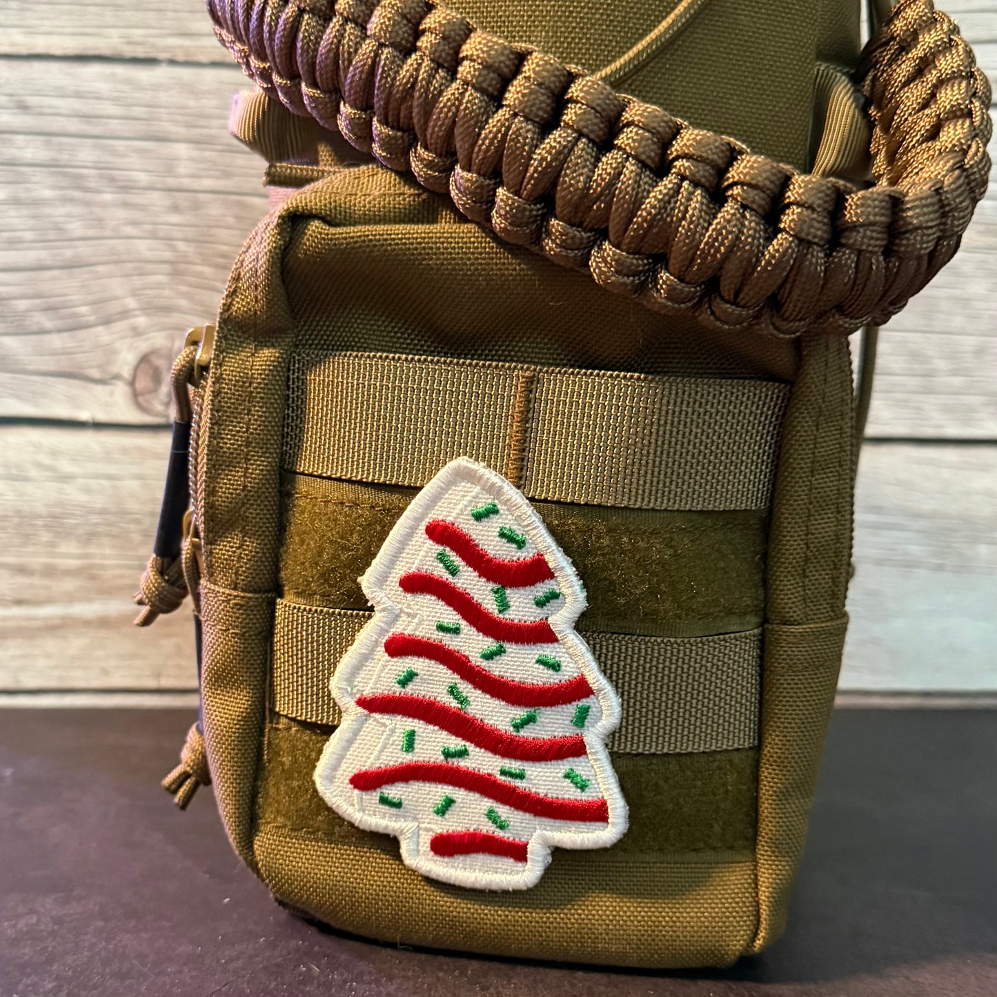 A close-up of a tan tactical pouch with a thick braided brown strap and a white embroidered Christmas tree patch with red and green stripes, set against a light wood plank background.