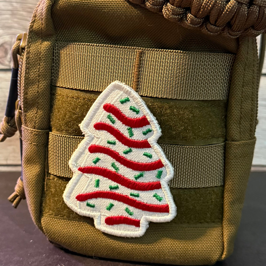 A close-up of a tan tactical pouch with a white embroidered Christmas tree patch featuring red stripes and green accents, attached to the front webbing.