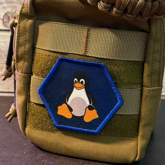 A tactical pouch in olive drab green nylon with a thick brown paracord loop handle, featuring a hexagonal blue patch with a penguin design on the front, set against a rustic wooden background.