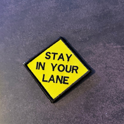A diamond-shaped yellow embroidered patch with a black border and the text 'STAY IN YOUR LANE' in black capital letters, placed on a dark textured gray surface.