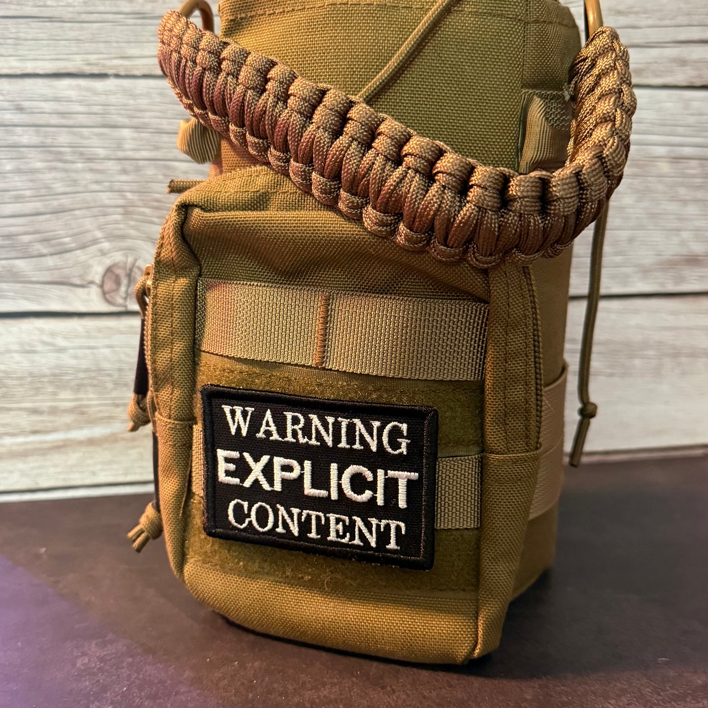 A tan tactical pouch with a brown paracord carrying strap, featuring a black rectangular patch with the text 'WARNING EXPLICIT CONTENT' in white. The pouch is set against a rustic gray wood plank background.