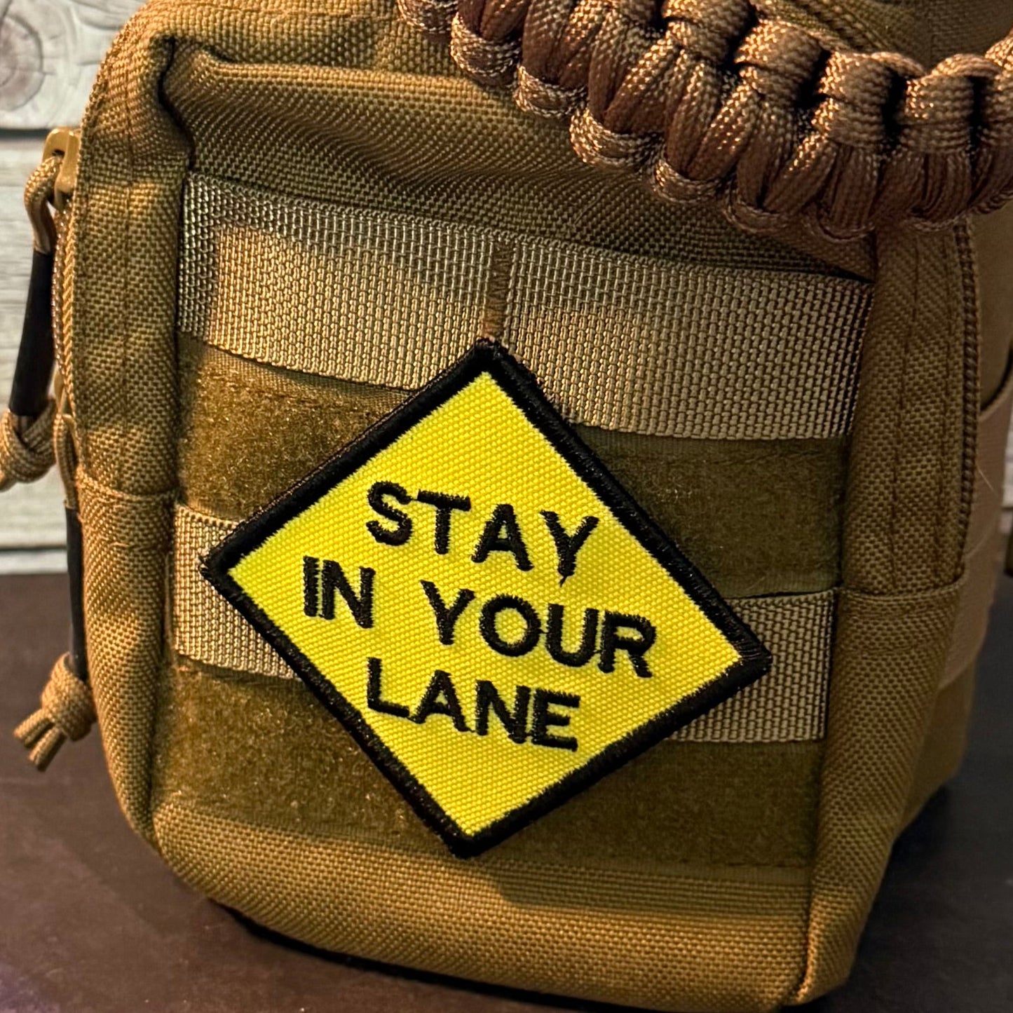 A tan tactical utility pouch with a paracord shoulder strap and a yellow diamond-shaped patch that reads 'STAY IN YOUR LANE'.