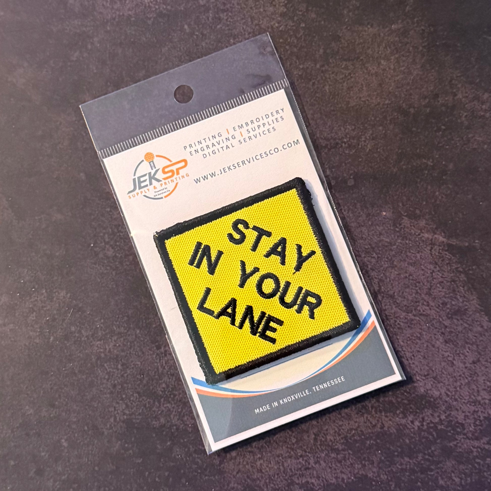 A yellow rectangular embroidered patch with black border and the words 'STAY IN YOUR LANE' in black lettering, attached to a white backing card with the JEKSP logo, photographed on a dark textured surface.