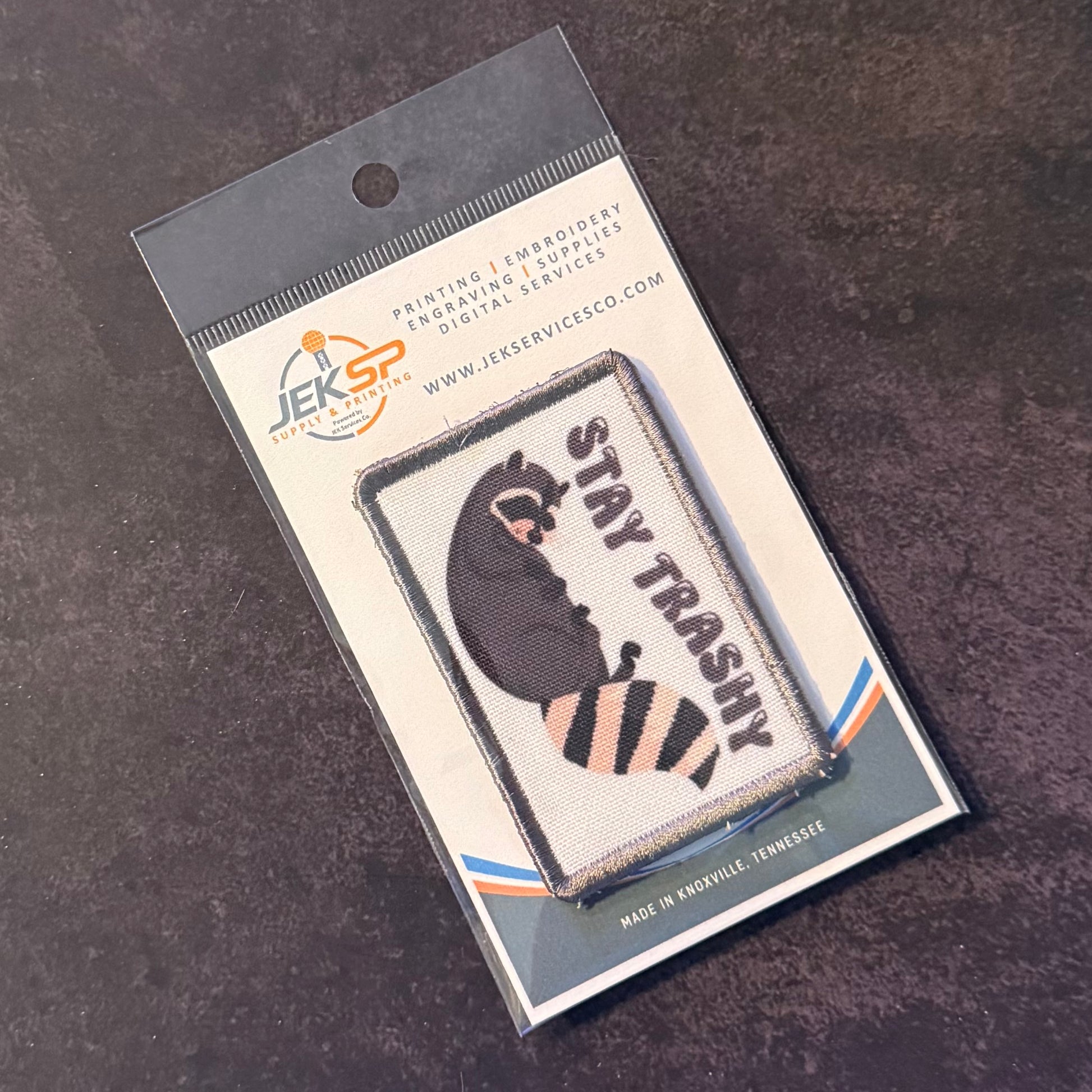 An embroidered patch featuring a raccoon with the text 'STAY TRASHY' in black and orange, packaged on a branded backing card from JEKSP Embroidery Services, photographed on a dark textured surface.