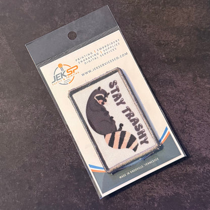 An embroidered patch featuring a raccoon with the text 'STAY TRASHY' in black and orange, packaged on a branded backing card from JEKSP Embroidery Services, photographed on a dark textured surface.