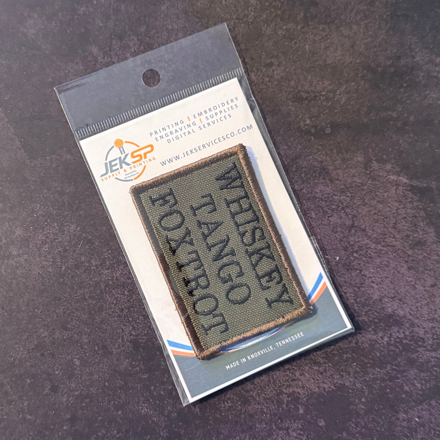 A rectangular embroidered patch reading 'WHISKEY TANGO ROXTRON' in dark lettering on an olive drab background, mounted on a printed backing card and photographed on a dark textured surface.