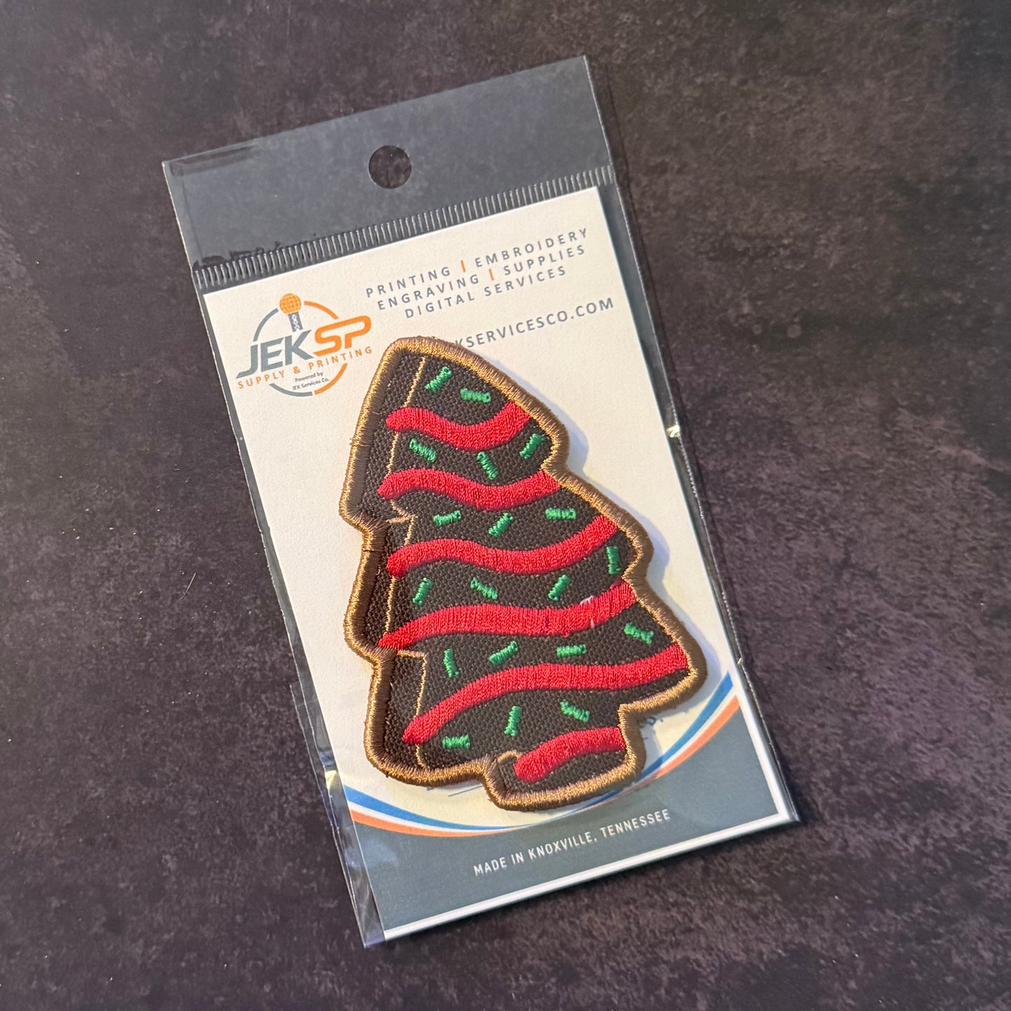 An embroidered Christmas tree patch in a clear plastic hanging sleeve with a printed backing card. The patch is dark brown with red wavy stripes and small green accents, and the card shows the JEKSP brand and text including 'PRINTING | EMBROIDERY | ENGRAVING | SUPPLIES' and 'SERVICESCO.