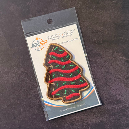 An embroidered Christmas tree patch in a clear plastic hanging sleeve with a printed backing card. The patch is dark brown with red wavy stripes and small green accents, and the card shows the JEKSP brand and text including 'PRINTING | EMBROIDERY | ENGRAVING | SUPPLIES' and 'SERVICESCO.