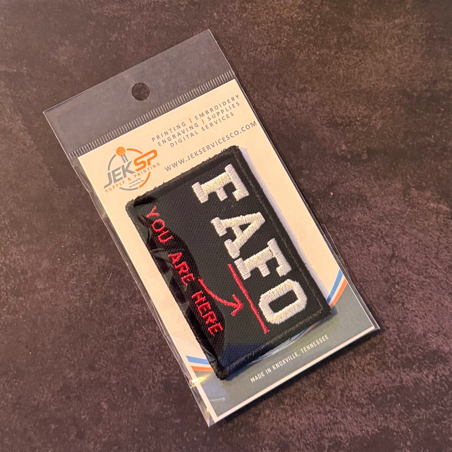 A black rectangular embroidered patch with white 'EAFO' lettering and red 'YOU ARE HERE' text, sealed in clear retail packaging with a printed backing card, photographed on a dark textured surface.