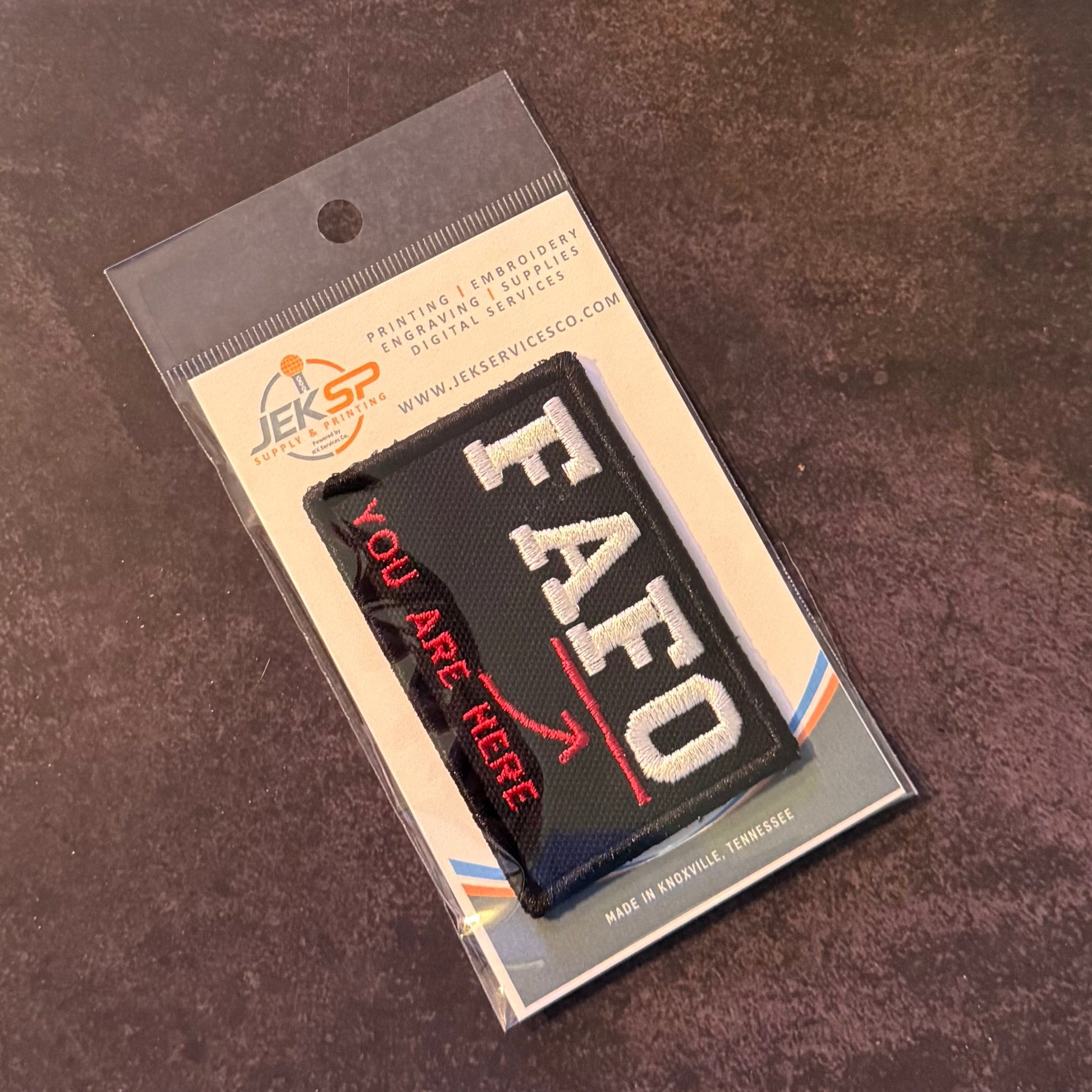 A black rectangular embroidered patch with white 'EAFO' lettering and red 'YOU ARE HERE' text, sealed in clear retail packaging with a printed backing card, photographed on a dark textured surface.