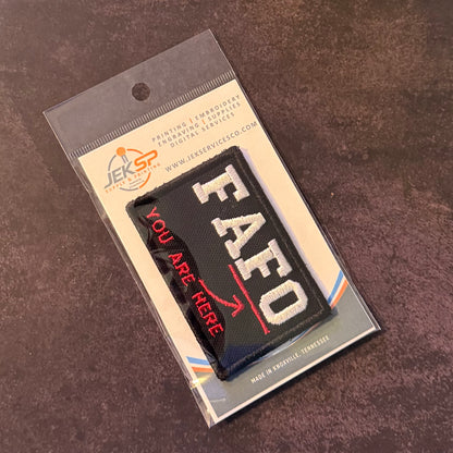 A black rectangular embroidered patch with white 'EAFO' lettering and red 'YOU ARE HERE' text, sealed in clear retail packaging with a printed backing card, photographed on a dark textured surface.