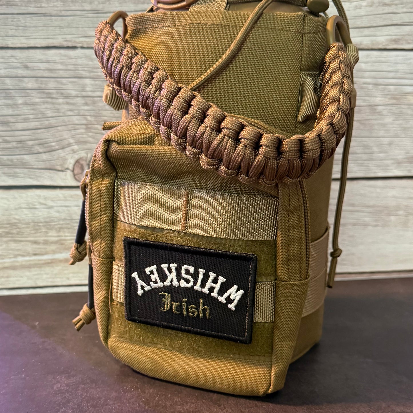 A tan tactical pouch bag with a brown paracord shoulder strap, featuring a black rectangular patch with white and gold text that reads 'MEXICO Irish'. The pouch is set against a rustic light gray wood plank background.