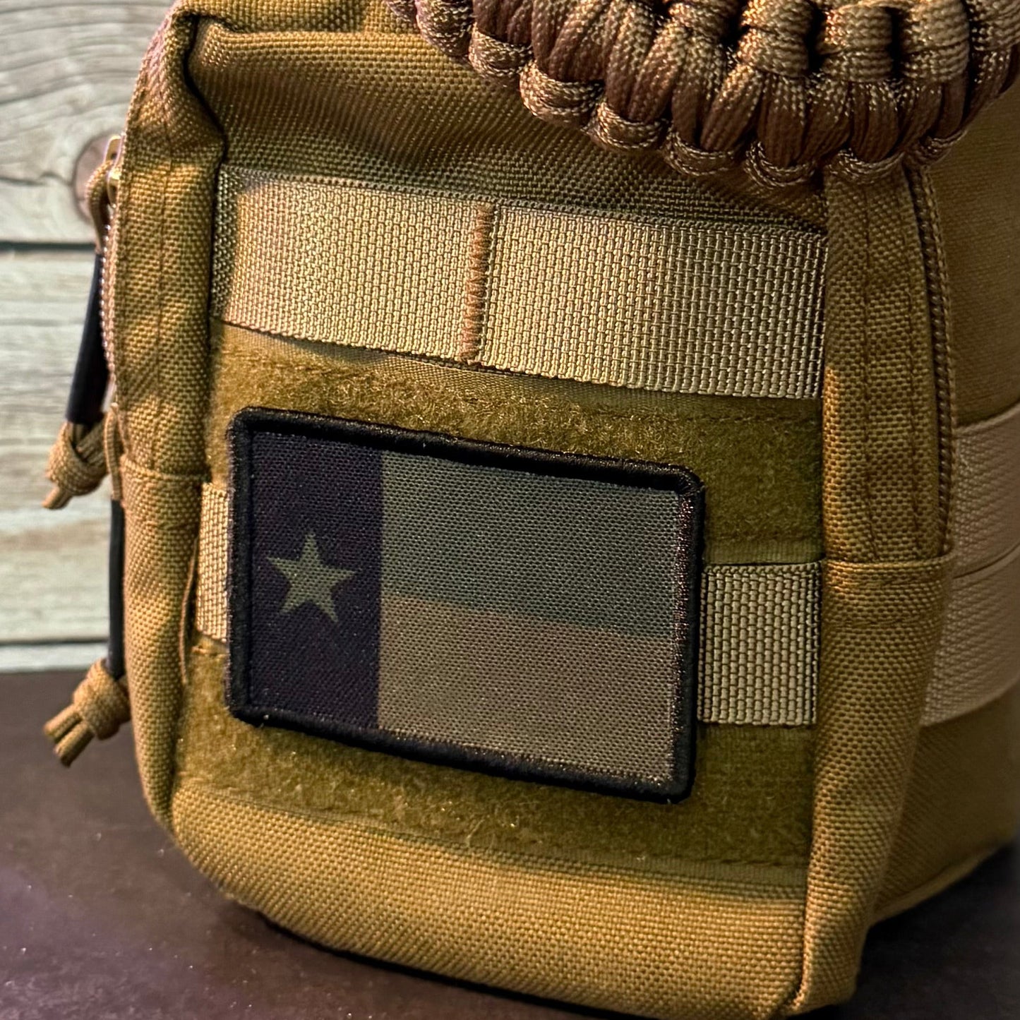 A tactical utility pouch in coyote brown and tan canvas, featuring a woven paracord shoulder strap, a front zip pocket with a Texas flag patch, and a wooden plank background.