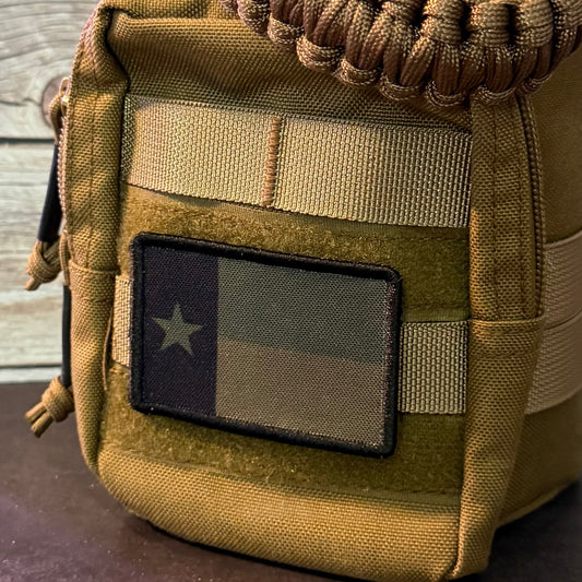 A tactical utility pouch in coyote brown and tan canvas, featuring a woven paracord shoulder strap, a front zip pocket with a Texas flag patch, and a wooden plank background.
