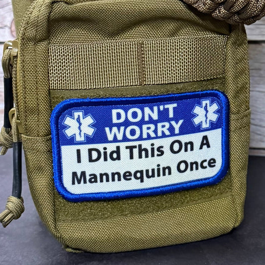 An olive green tactical pouch with a paracord strap, featuring a blue and white patch that reads 'DON'T WORRY I Did This On A Mannequin Once' with Star of Life icons.