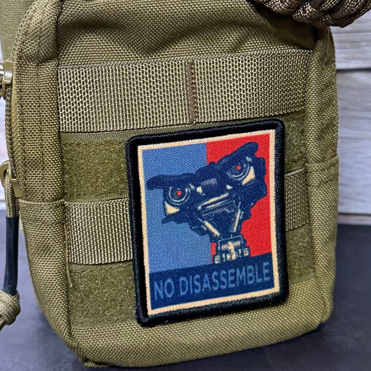 An olive green tactical pouch with a woven brown strap, featuring a rectangular patch with a stylized robot face and the text 'NO DISASSEMBLE' in blue on a red and blue background.