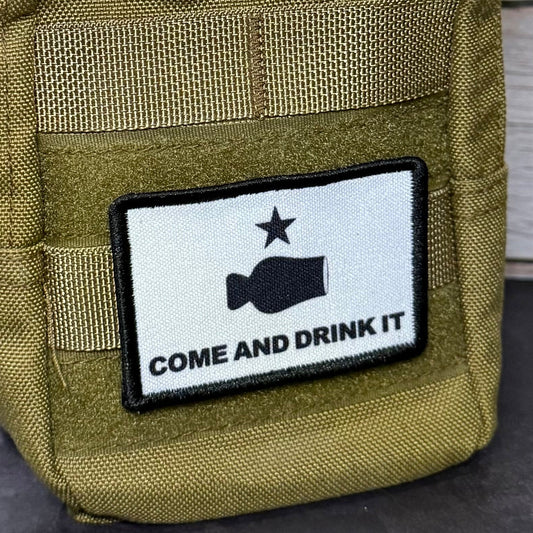 An olive drab tactical pouch with a paracord strap, featuring a white rectangular patch with a black fish graphic and the text 'COME AND DRINK IT'.