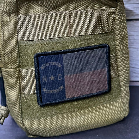 An olive drab tactical pouch with a rectangular embroidered patch featuring a dark blue, black, and brown vertical tricolor design with 'N C' in the center, shown against a light wood plank background.