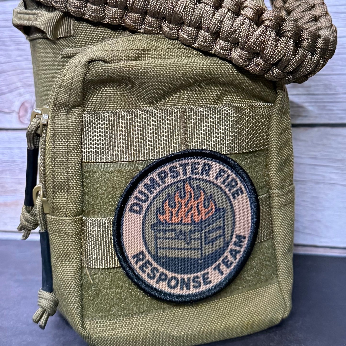 An olive drab tactical utility pouch with a woven paracord shoulder strap, featuring a circular 'Dumpster Fire Response Team' patch on the front, set against a light gray wood plank background.