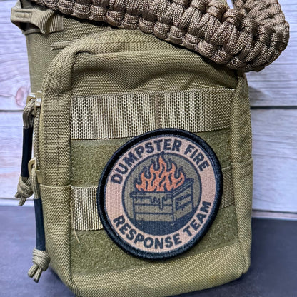 An olive drab tactical utility pouch with a woven paracord shoulder strap, featuring a circular 'Dumpster Fire Response Team' patch on the front, set against a light gray wood plank background.
