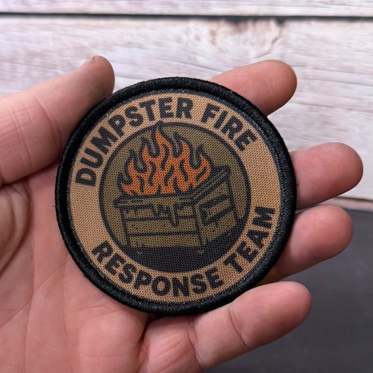 A round embroidered patch reading 'DUMPSTER FIRE RESPONSE TEAM' with a fire graphic, held in a person's hand against a light wood and dark gray background.