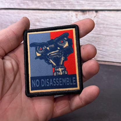 A hand holding a square embroidered patch featuring a stylized robot head over a split blue and red background with the text 'NO DISASSEMBLE' at the bottom.