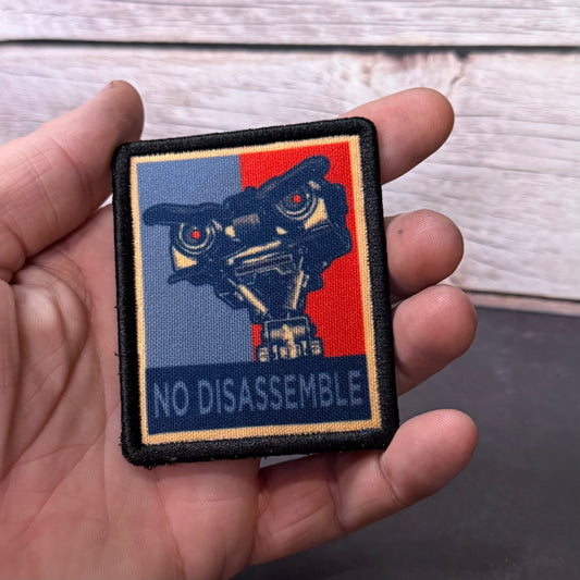 A hand holding a square embroidered patch featuring a stylized robot head over a split blue and red background with the text 'NO DISASSEMBLE' at the bottom.