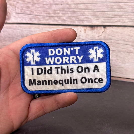 A hand holding a rectangular embroidered patch with a blue border and white center. The patch has a blue top section with white Star of Life symbols and the words 'DON'T WORRY,' and a white bottom section with the phrase 'I Did This On A Mannequin Once' in black text.