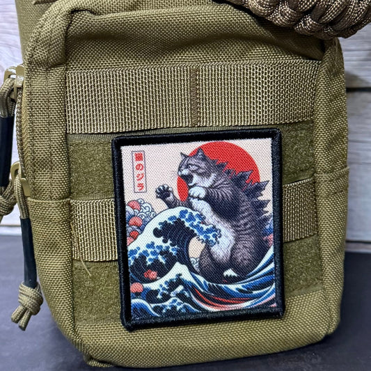 An olive green tactical pouch with a woven paracord strap, featuring a rectangular patch with a Japanese-style cat wave artwork, set against a light wood background.