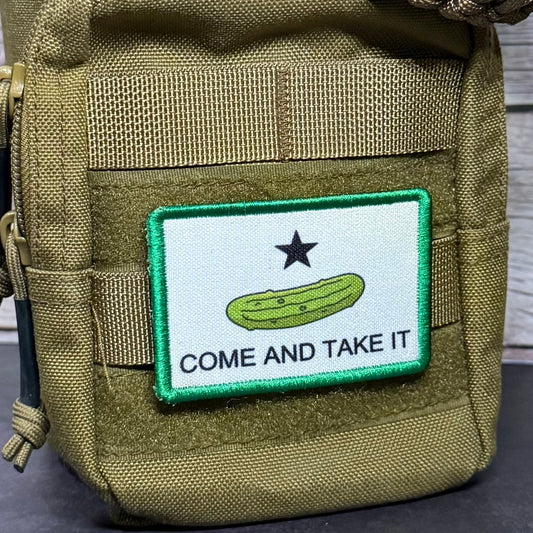Rectangular Come and Take It morale patch with a green pickle, black star, black lettering, and green border attached to an olive tactical pouch.