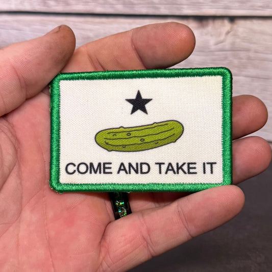 A hand holding a rectangular embroidered patch featuring a white field with a green border, a black star, a green pickle illustration, and the text 'COME AND TAKE IT' in black. The background shows a light wood surface and dark gray floor.