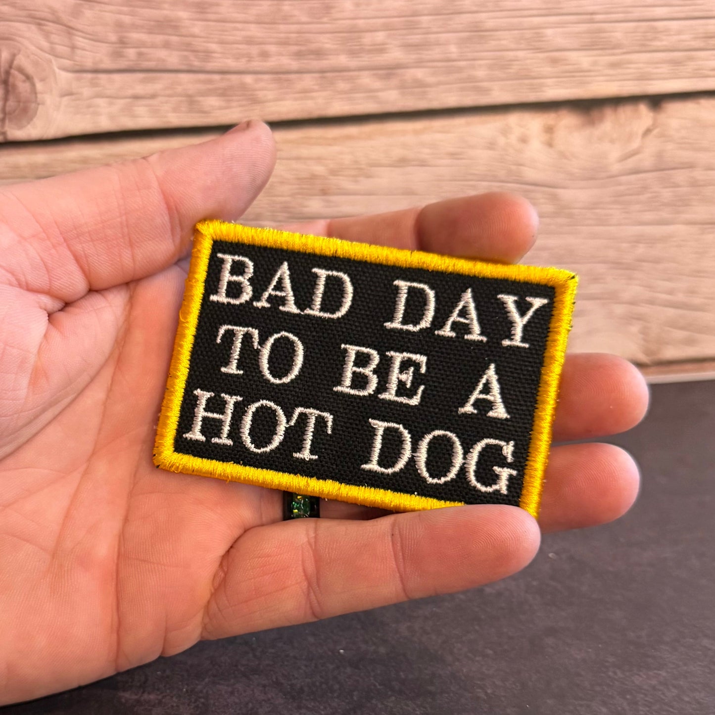 A hand holding a rectangular embroidered patch with the text 'BAD DAY TO BE A HOT DOG' in white on a black background with a yellow border, set against a rustic wooden background.