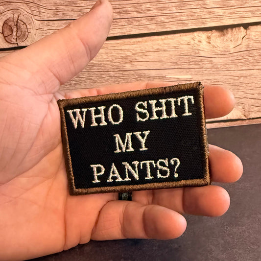 A rectangular embroidered patch held in a hand, featuring the text 'WHO SHIT MY PANTS?' in white lettering on a black background with a tan border, set against a rustic wooden plank backdrop.