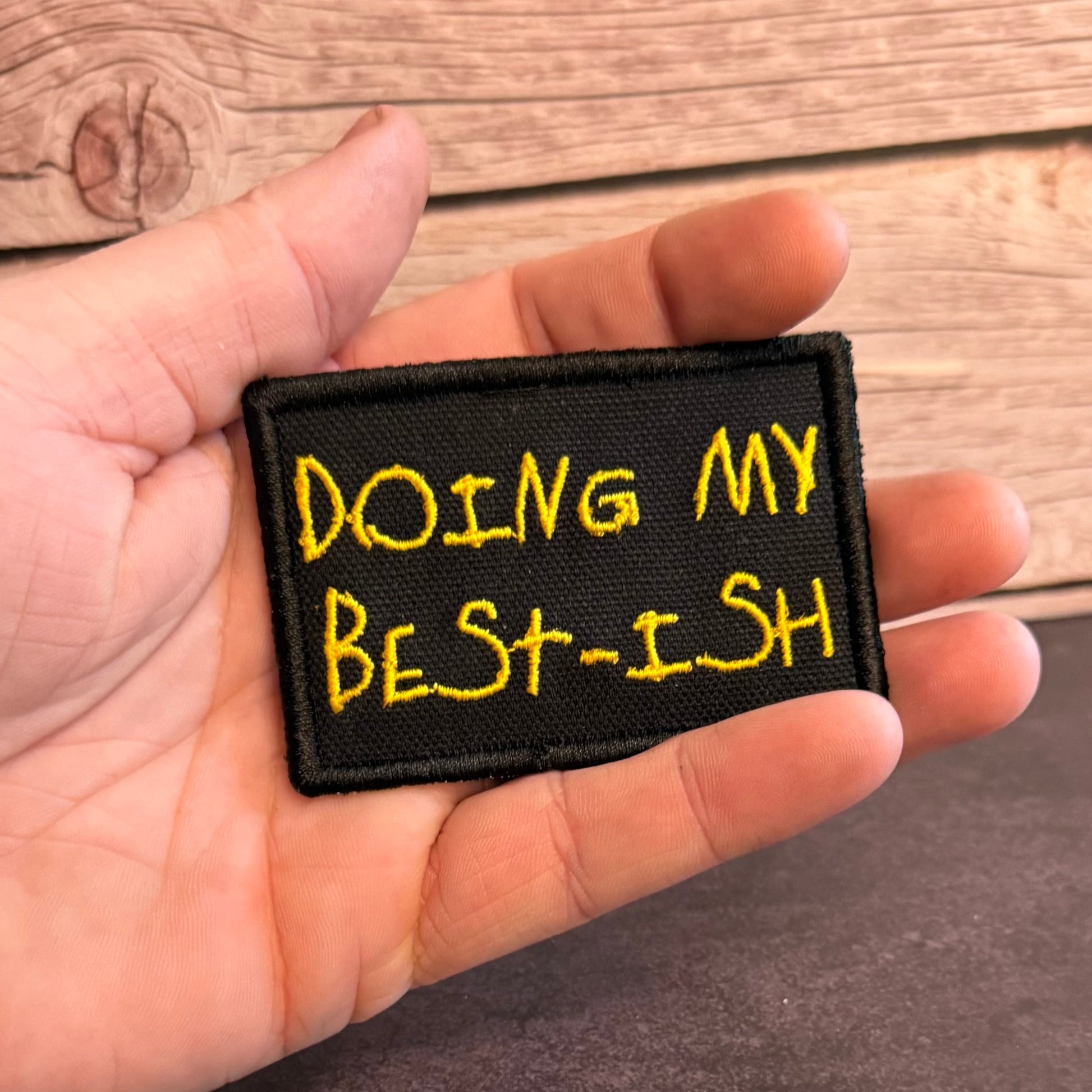 A hand holding a rectangular black embroidered patch with yellow text that reads 'DOING MY BEST-ISH' against a rustic wood background.