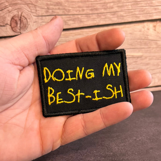 A hand holding a rectangular black embroidered patch with yellow text that reads 'DOING MY BEST-ISH' against a rustic wood background.