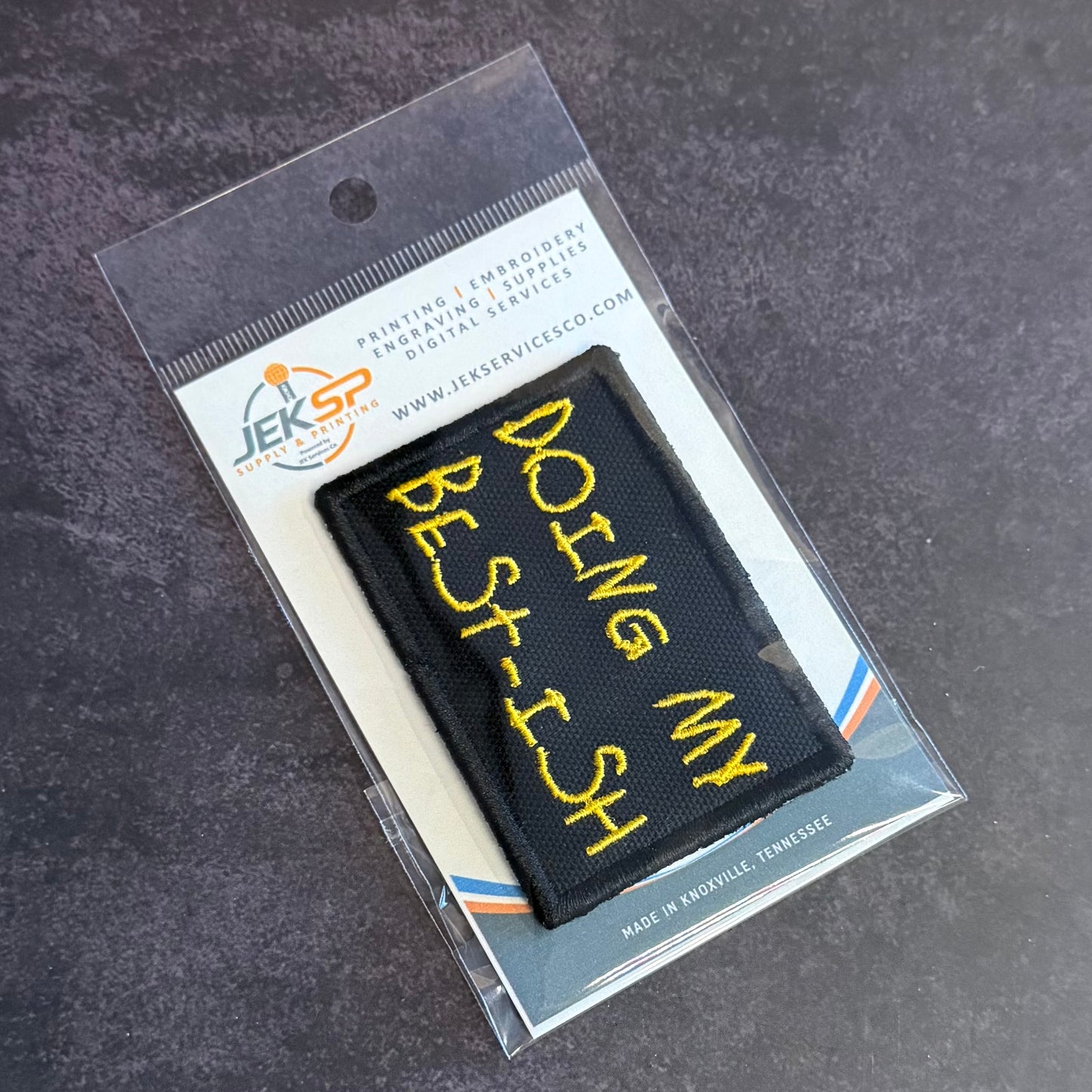 A black rectangular embroidered patch with yellow text reading 'DOING MY BEST ISH' inside a clear plastic retail sleeve with a printed backing card. The JEKSP brand logo is visible on the card.
