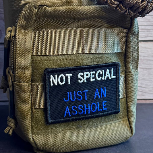 An olive drab tactical pouch with a black embroidered patch that reads 'NOT SPECIAL JUST AN ASSHOLE' in white and blue text, shown with a paracord rope handle against a rustic wood background.
