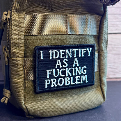A close-up of a tactical pouch in olive drab/khaki nylon with a black rectangular embroidered patch on the front that reads 'I IDENTIFY AS A FUCKING PROBLEM' in white text.