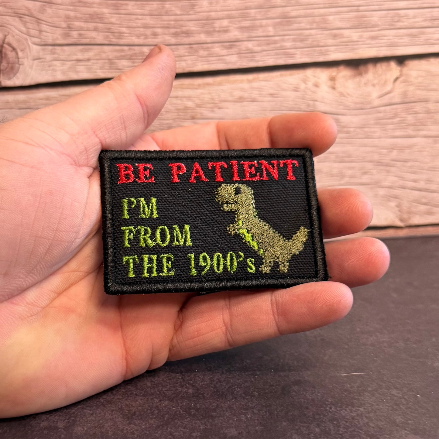 A hand holding a rectangular embroidered patch with a black background, red text reading 'BE PATIENT', yellow text reading 'PM FROM THE 1900's', and a yellow dinosaur silhouette.