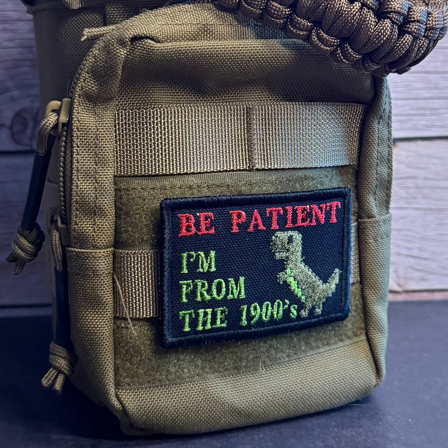 An olive green tactical pouch with a paracord strap, featuring a custom embroidered patch that reads 'BE PATIENT I'M FROM THE 1900's' with a dinosaur graphic.