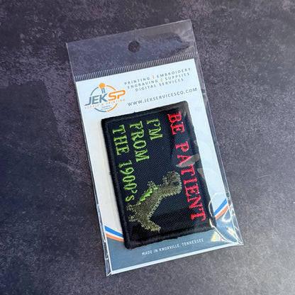 A JEKSP embroidered patch in its clear plastic retail packaging, featuring a black background with green and red text reading 'I'M FROM THE 1990's BE PATIENT' and a green dinosaur graphic.