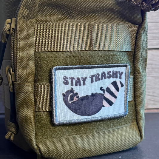 An olive green tactical pouch with a front hook-and-loop patch that reads 'STAY TRASHY' and shows a raccoon illustration, photographed against a wood-paneled background.