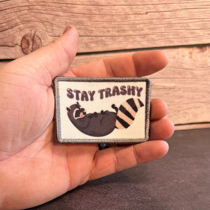 A hand holding a rectangular embroidered patch that reads “STAY TRASHY” above a sleeping raccoon with a striped tail, set against a rustic wood plank background.