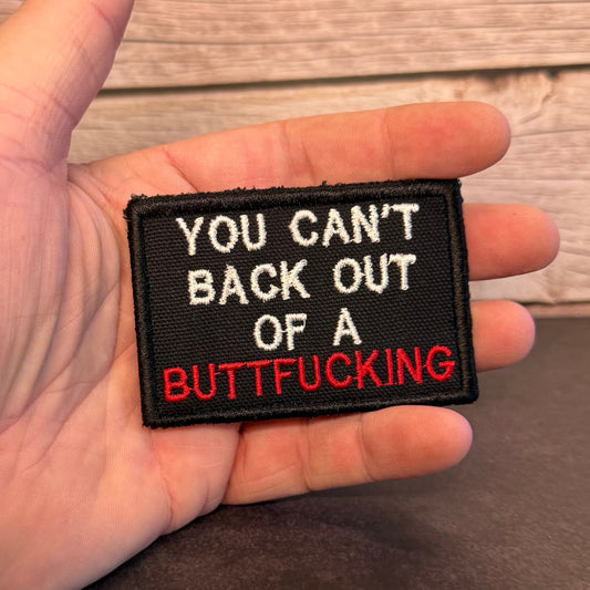 A rectangular embroidered patch held in a hand, featuring the text 'YOU CAN'T BACK OUT OF A BUTTFUCKING' in white and red lettering on a black background, with a wood plank wall behind it.